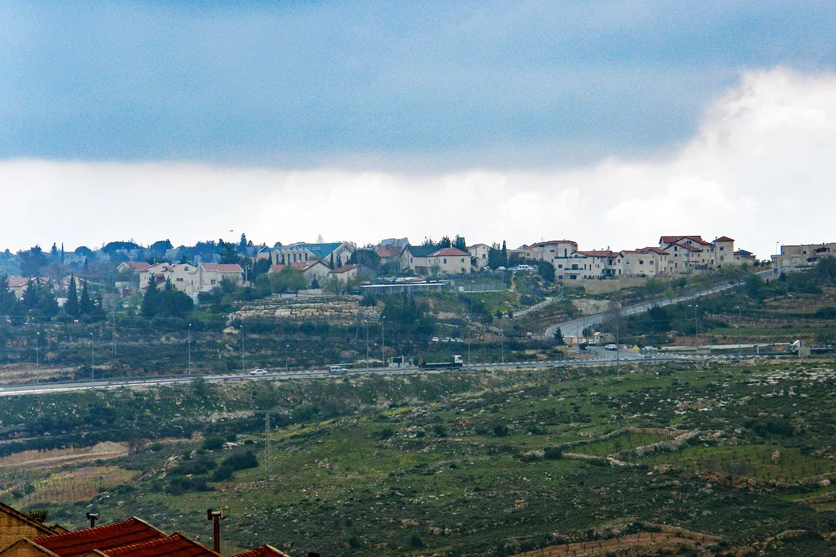 Elazar's houses along a ridge above a main road and green fields, under a cloudy sky